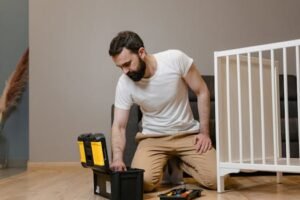 A man kneels indoors assembling a baby crib using a toolbox, wearing a white shirt and beige pants.